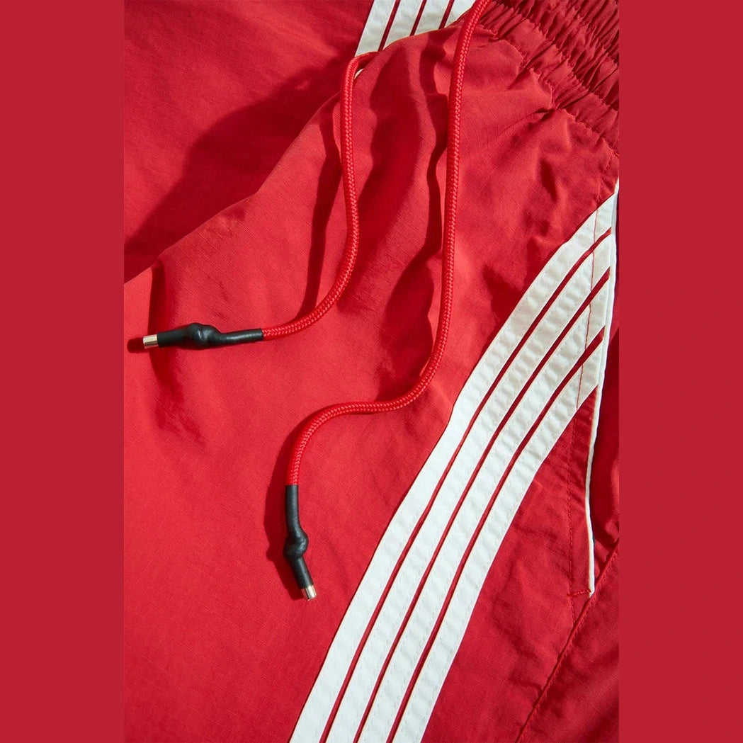Red Jordan Craig The Beautiful Game track pants featuring white side stripes, elastic waistband, and red drawstrings with black aglets.